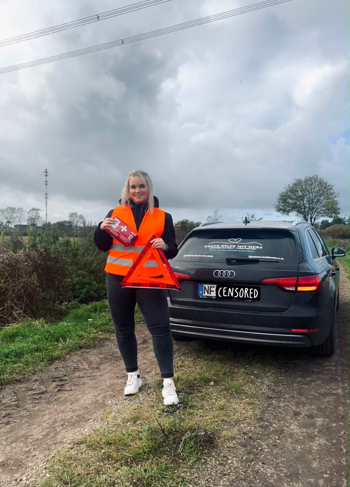 Jana Marie Paulsen in orangefarbener Warnweste mit Warndreieck neben einem schwarzen Audi auf dem Land.