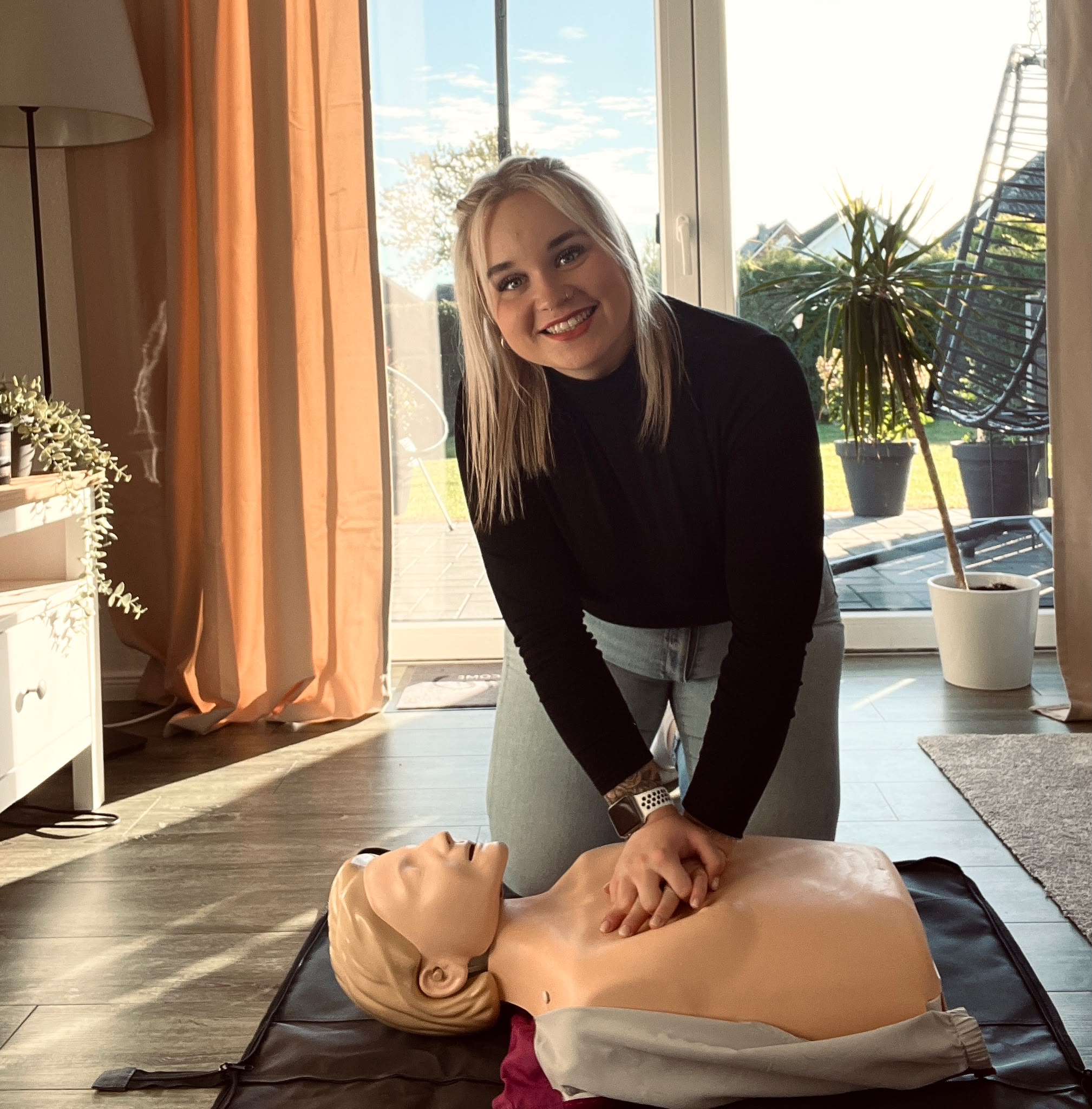 Jana Marie Paulsen lächelt in die Kamera, während sie eine Herzdruckmassage an einer CPR-Trainingspuppe demonstriert.