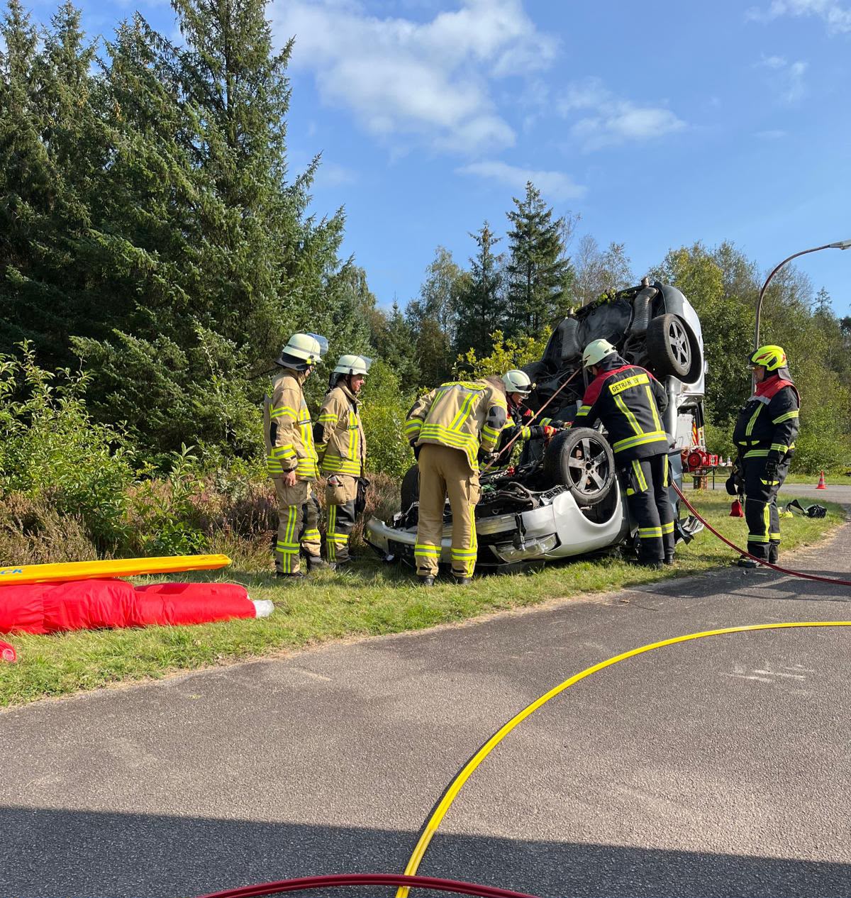 Feuerwehrleute in voller Einsatzmontur sichern ein auf dem Dach liegendes Unfallfahrzeug am Straßenrand.